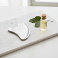 AVELUNE Sculpting Gua Sha stainless steel facial tool on marble surface with unlabeled glass bottle and green leaves